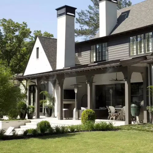A large residential home in Cherokee County featuring a covered backyard patio, where a fine mist from a mosquito control system is visibly spraying from the eaves over the outdoor living and seating area
