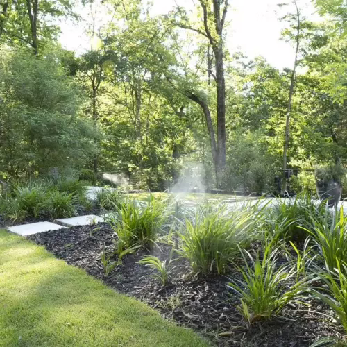 Automated nozzles spraying a fine mist over a landscaped garden bed next to a grassy lawn